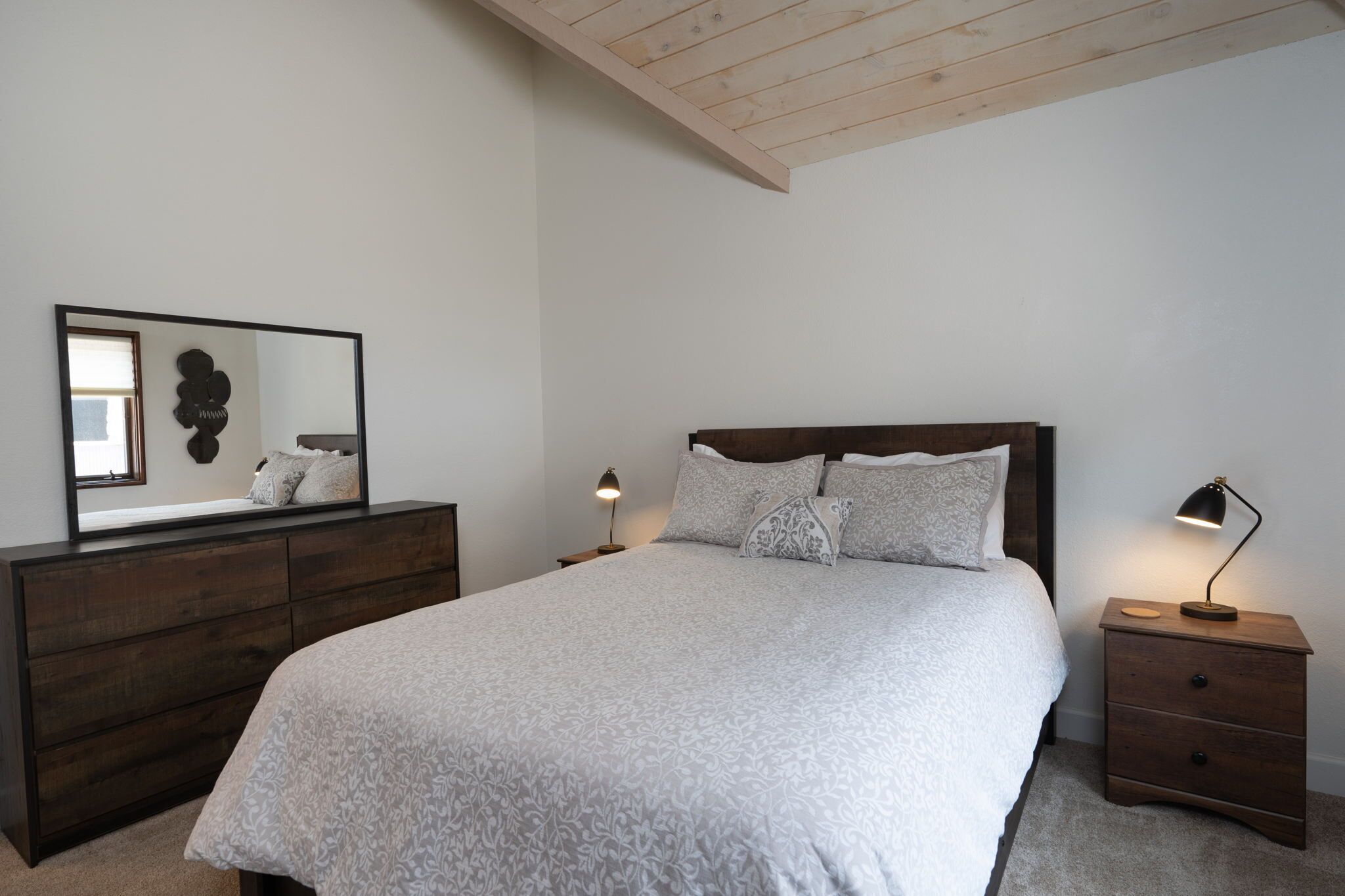 Un dormitorio con una cama grande, un aparador de madera, una mesita de noche con lámpara y un espejo montado en la pared.