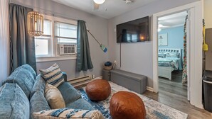 A modern living room with a grey sofa, leather ottomans, a ceiling fan, and a TV mounted on the wall.
