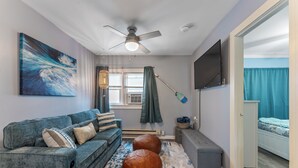 A modern living room with a grey sofa, two leather ottomans, a ceiling fan, a large abstract painting, and a television mounted on the wall.