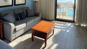 Ett rum med en grå soffa, ett träbord och utsikt över en pool och havet genom en stor skjutdörr i glas.