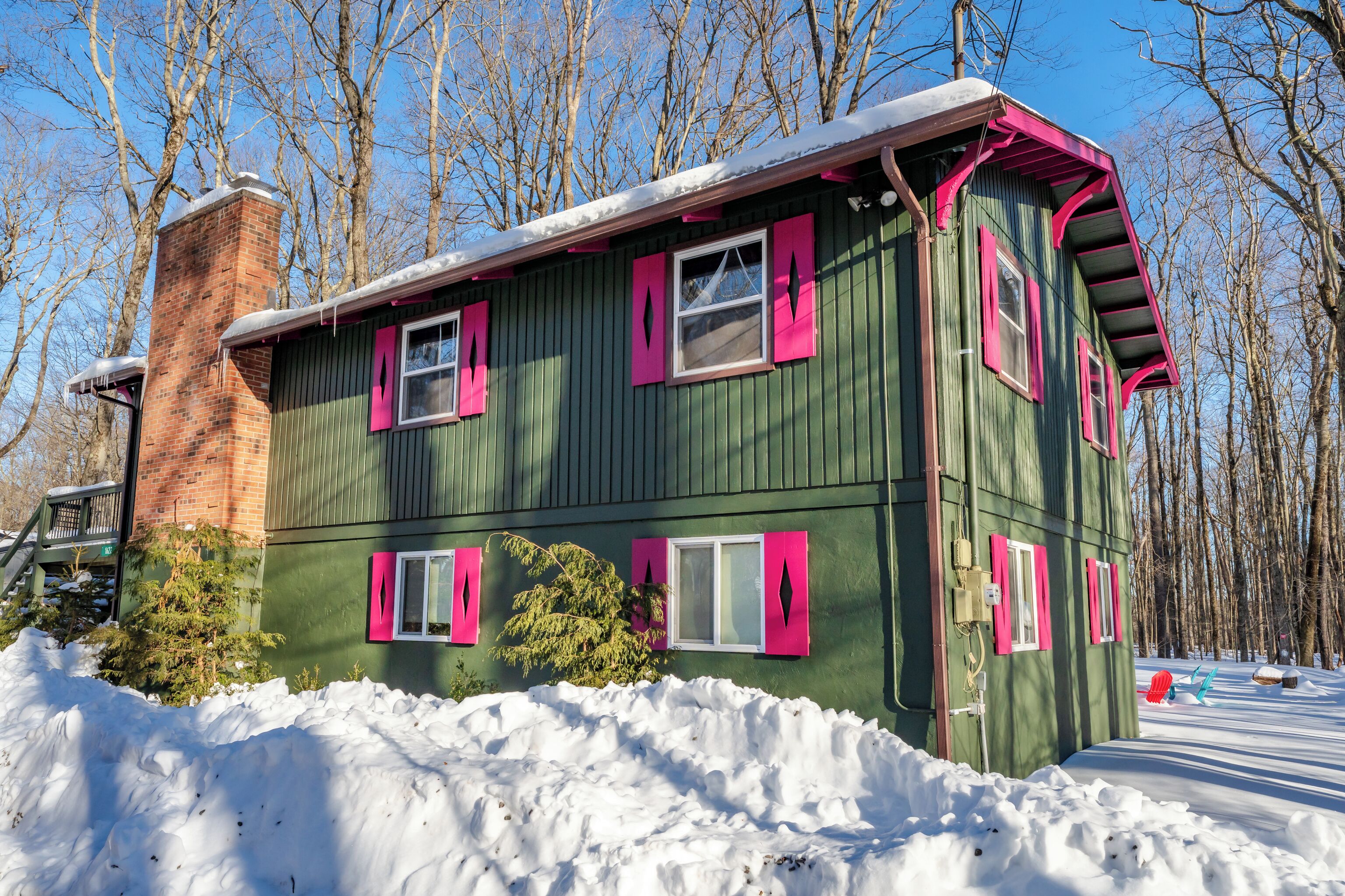 A green house with pink shutters and a brick chimney, surrounded by snow and trees.
