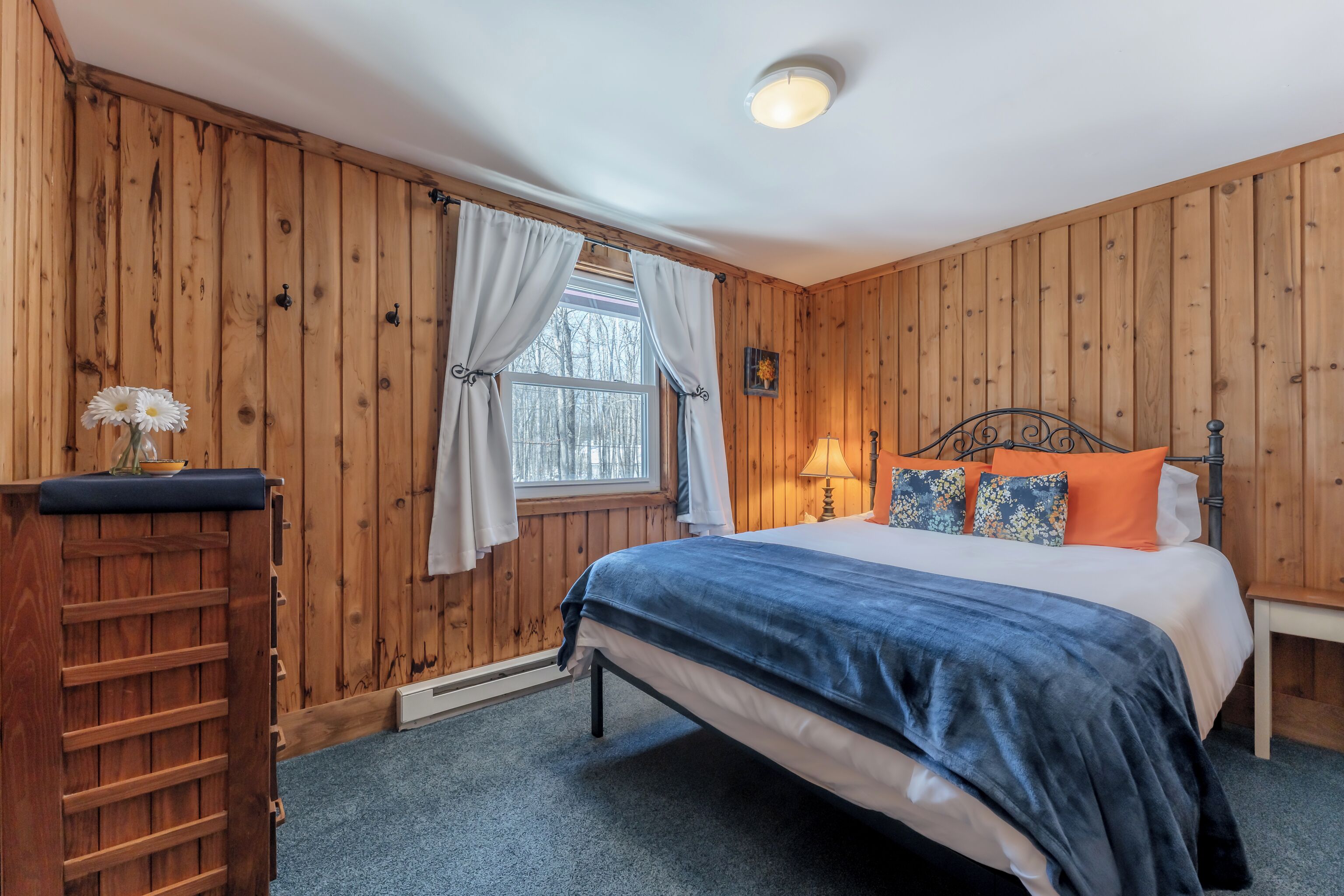 A bedroom with a wooden paneled wall, a bed with blue and white bedding, a nightstand, a lamp, and a window with curtains.