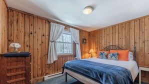 Ein Schlafzimmer mit einer holzverkleideten Wand, einem Bett mit blau-weißer Bettwäsche, einem Nachttisch, einer Lampe und einem Fenster mit Vorhängen.