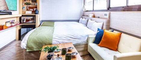 Habitación compacta con cama, sofá, mesa de madera con utensilios de cocina y ventana con vistas.