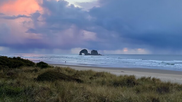 A beach with a natural rock formation in the distance, grassy dunes in the foreground, and a cloudy sky. - Peaceful Forest Cottage with Hot Tub  Near Beach+Twin Rocks  Family&Pet Friendly (Rockaway Beach)
