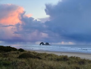 A beach with a natural rock formation in the distance, grassy dunes in the foreground, and a cloudy sky. - Peaceful Forest Cottage with Hot Tub  Near Beach+Twin Rocks  Family&Pet Friendly (Rockaway Beach)