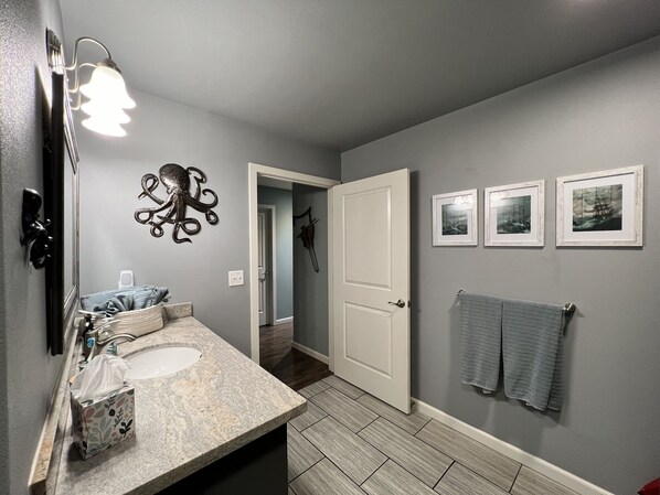 A bathroom with a granite countertop, a towel rack, and framed pictures on the wall. - Peaceful Forest Cottage with Hot Tub Near Beach+Twin Rocks Family&Pet Friendly (Rockaway Beach)