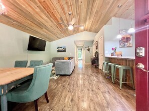 A spacious living area with a wooden ceiling, a dining table with chairs, a sofa, a TV mounted on the wall, and a kitchen area with stools. - Peaceful Forest Cottage with Hot Tub  Near Beach+Twin Rocks  Family&Pet Friendly (Rockaway Beach)