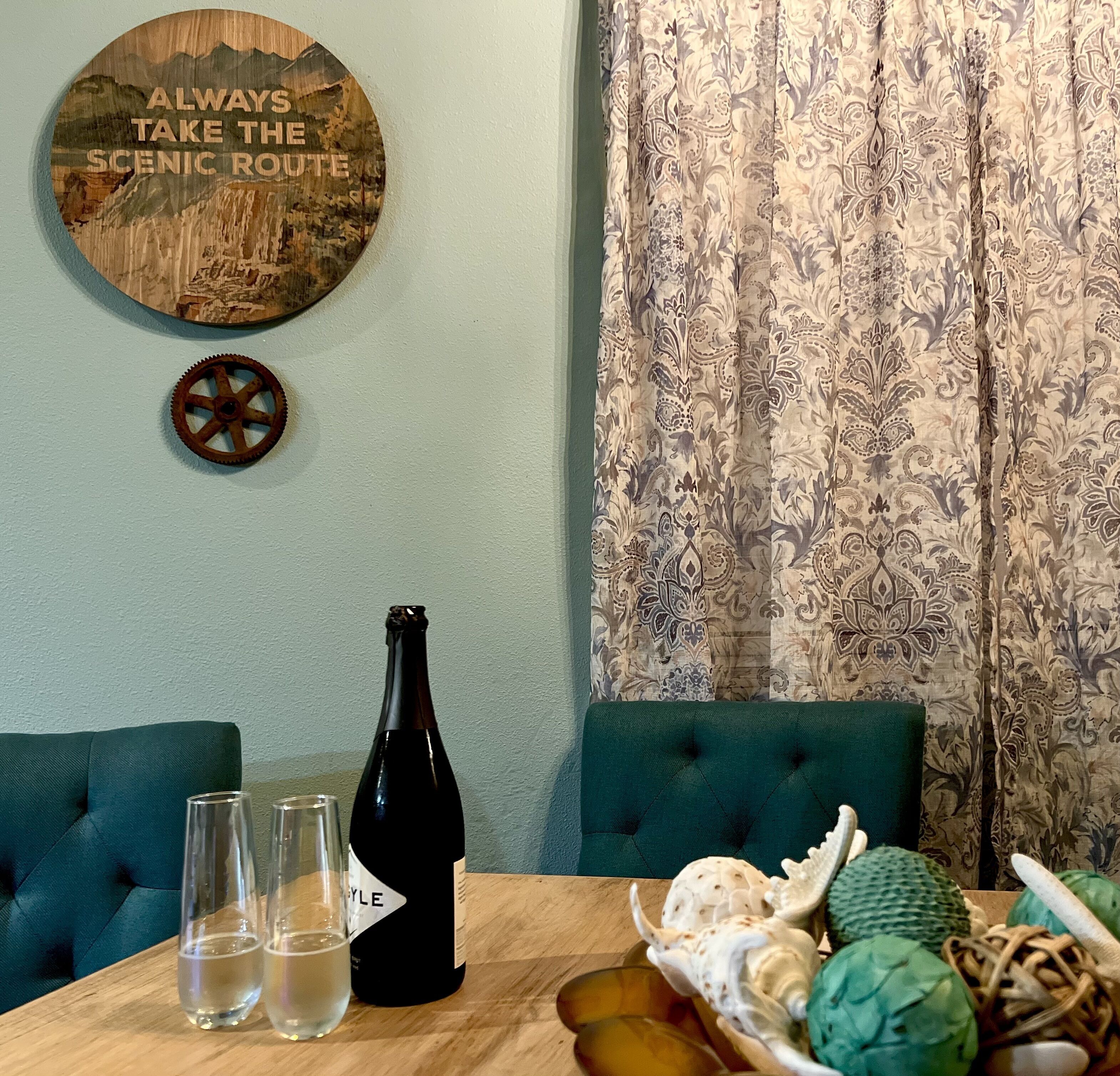 A dining table with a bottle of wine, two glasses, and a decorative plate with food. A wooden sign with a scenic route message is on the wall.