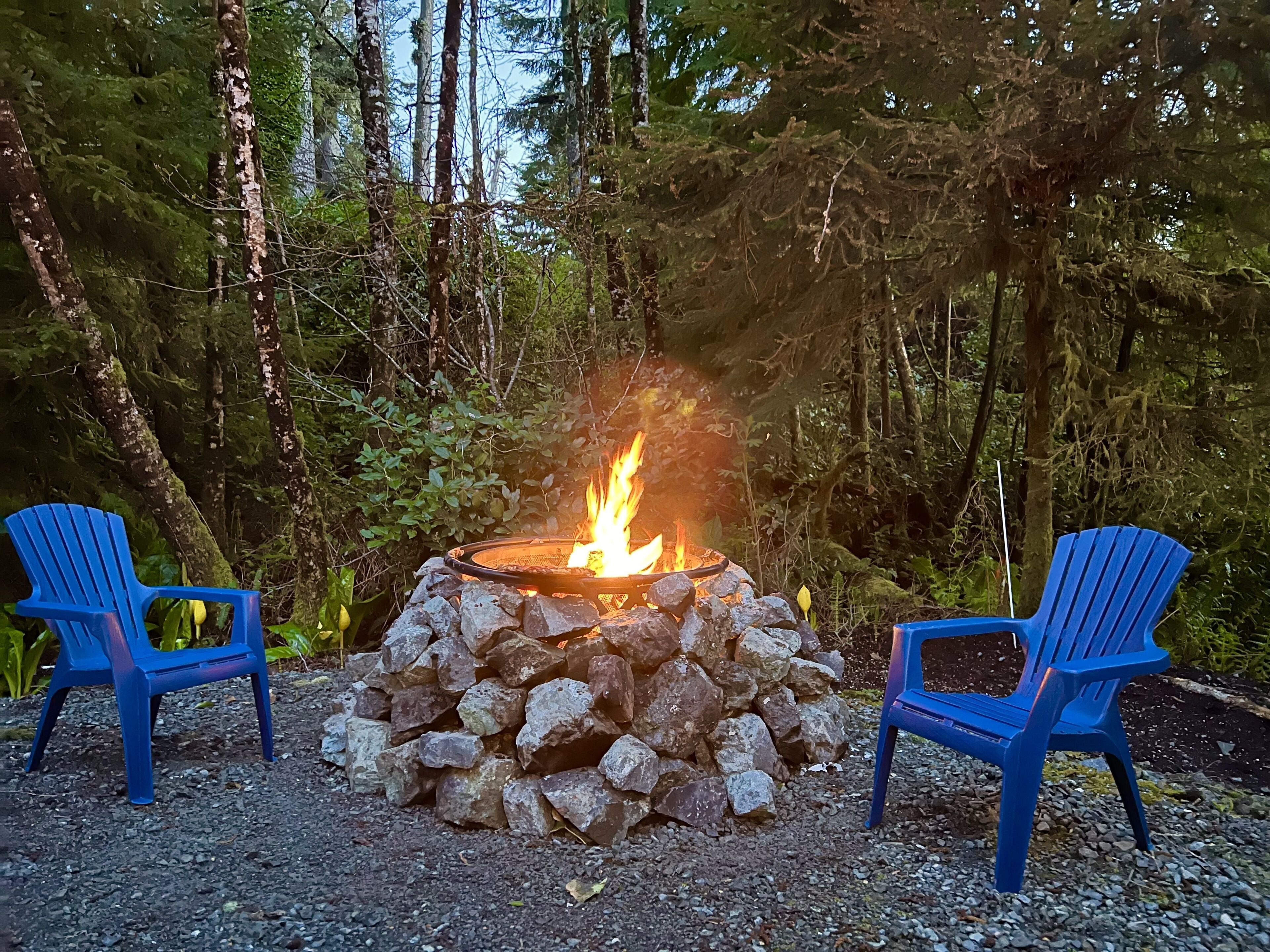 Two blue chairs facing a fire pit surrounded by rocks, set in a forested area.