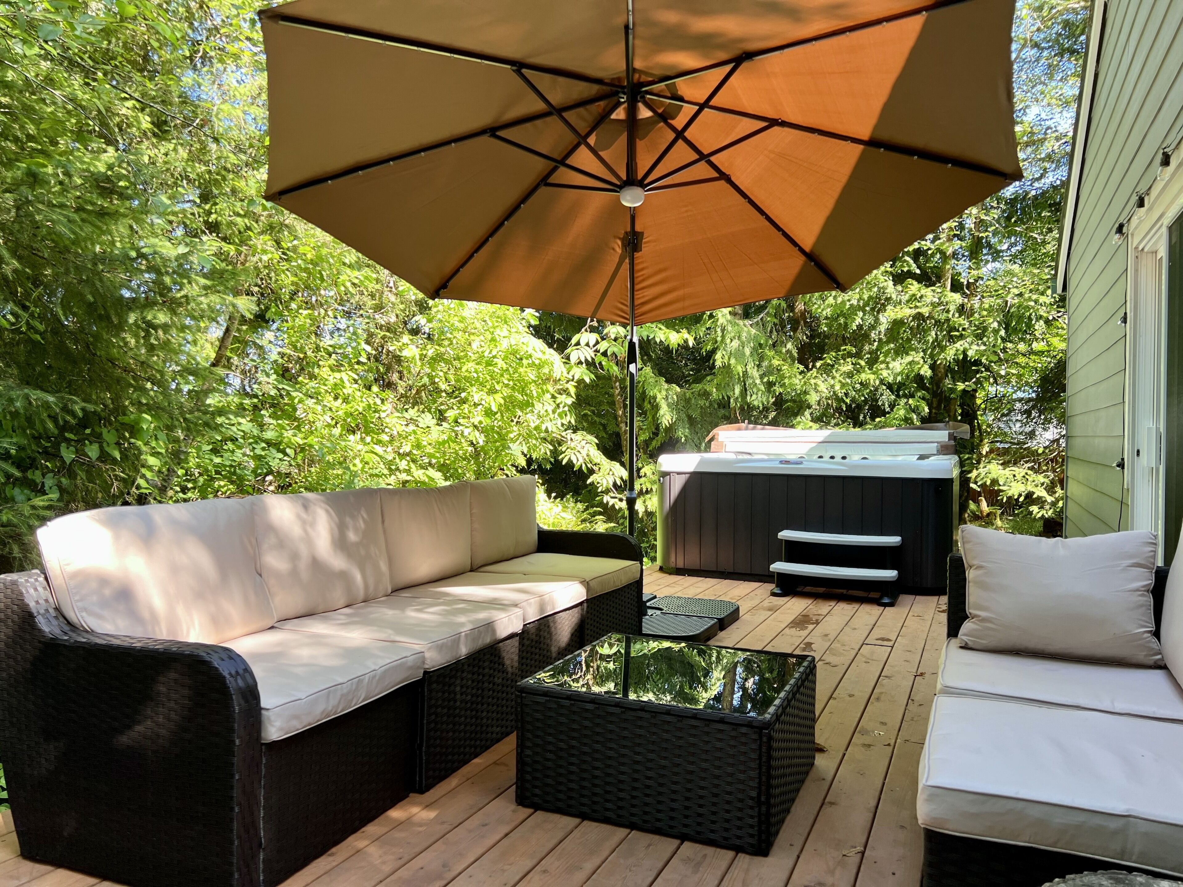 Eine Terrasse im Freien mit einem beigefarbenen Sofa, einem schwarzen Rattantisch und einem großen Sonnenschirm, der Schatten spendet.