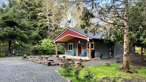 A small cabin with a red roof, two blue chairs outside, and a gravel driveway. - Peaceful Forest Cottage with Hot Tub  Near Beach+Twin Rocks  Family&Pet Friendly (Rockaway Beach)