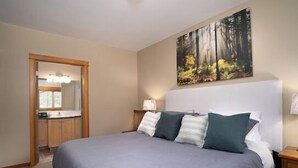 A bedroom with a large bed, a headboard, a painting of a forest, a nightstand, and a door leading to a bathroom. - Beautiful 2 bedroom condo in the heart of the Rocky Mountains (Canmore)