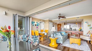 Un vaste espace de vie avec une porte-fenêtre coulissante donnant sur un balcon, un coin repas avec des chaises jaunes, une cuisine équipée d’appareils électroménagers en acier inoxydable et un ventilateur de plafond.