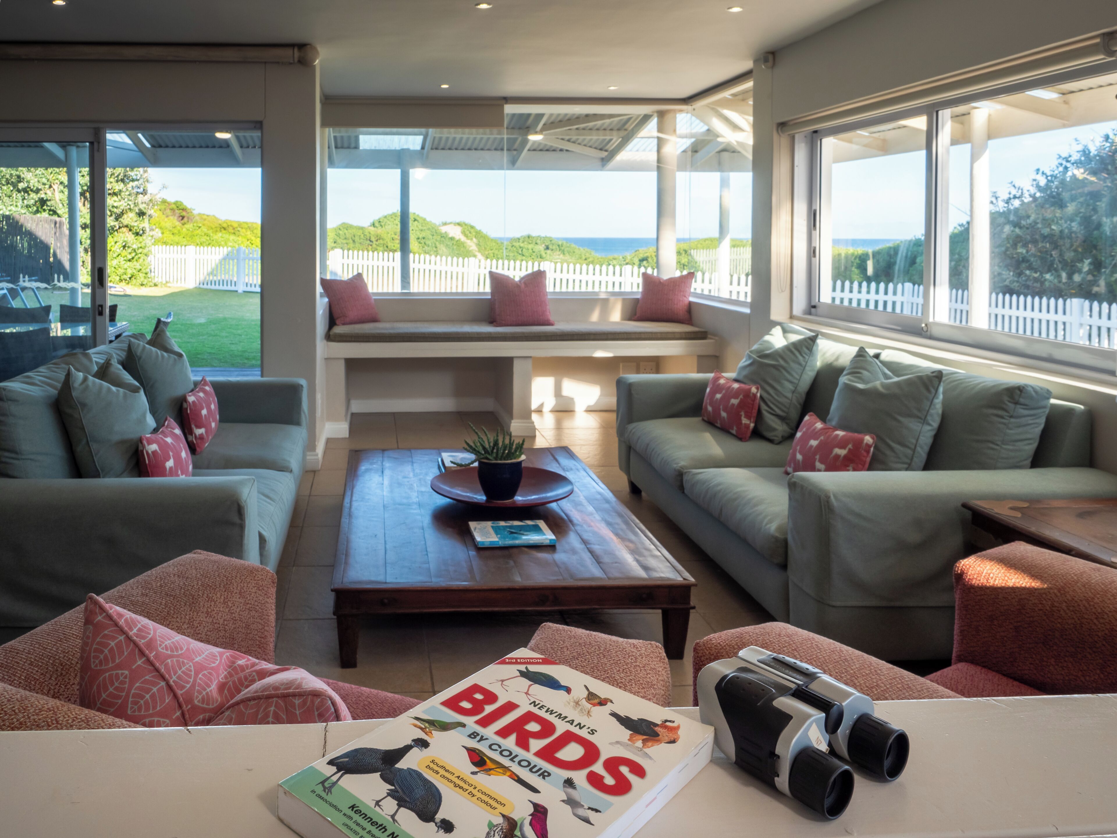 A modern living room with a large window, a sofa, a coffee table, and a book titled 'Birds by Colour'.