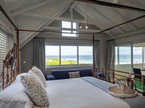 Ein Bett mit Baldachin und Blick auf den Ozean, ein Balkon mit Tisch sowie eine Champagnerflasche auf dem Bett.