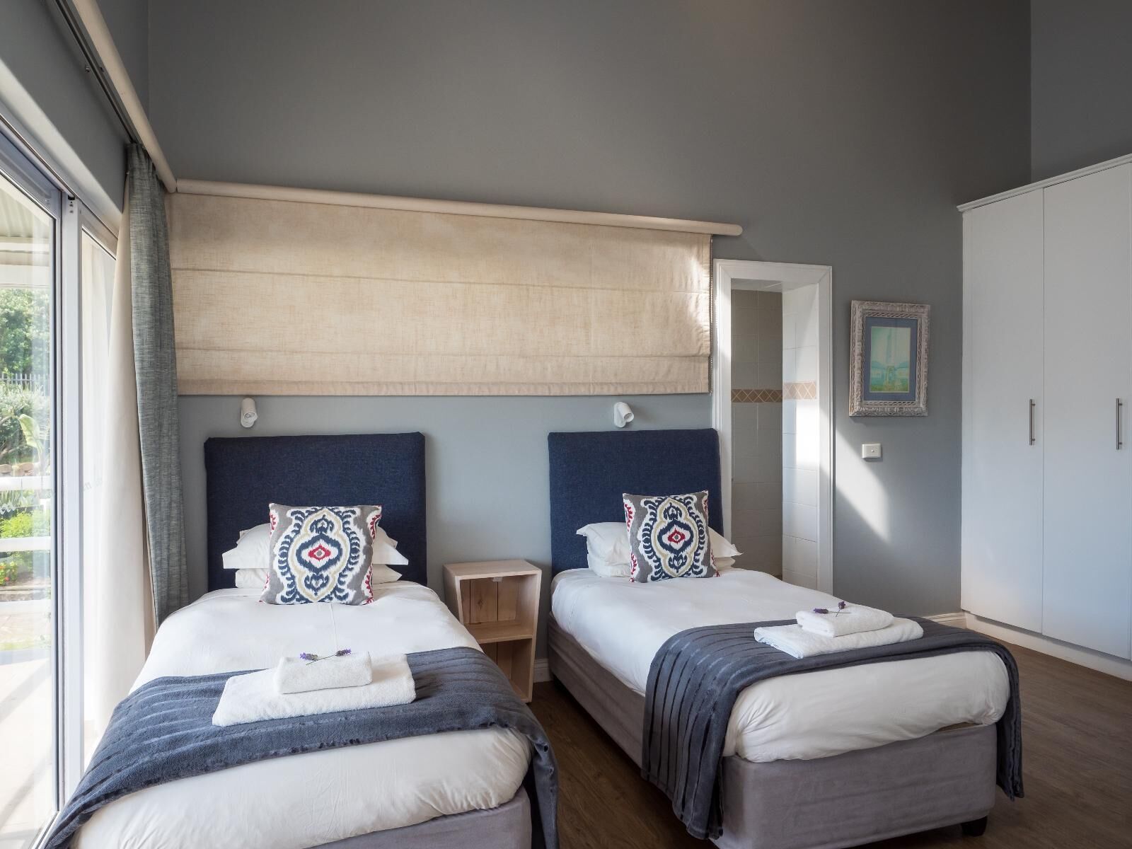 A hotel room with two beds, a large window, a wardrobe, and a painting on the wall.