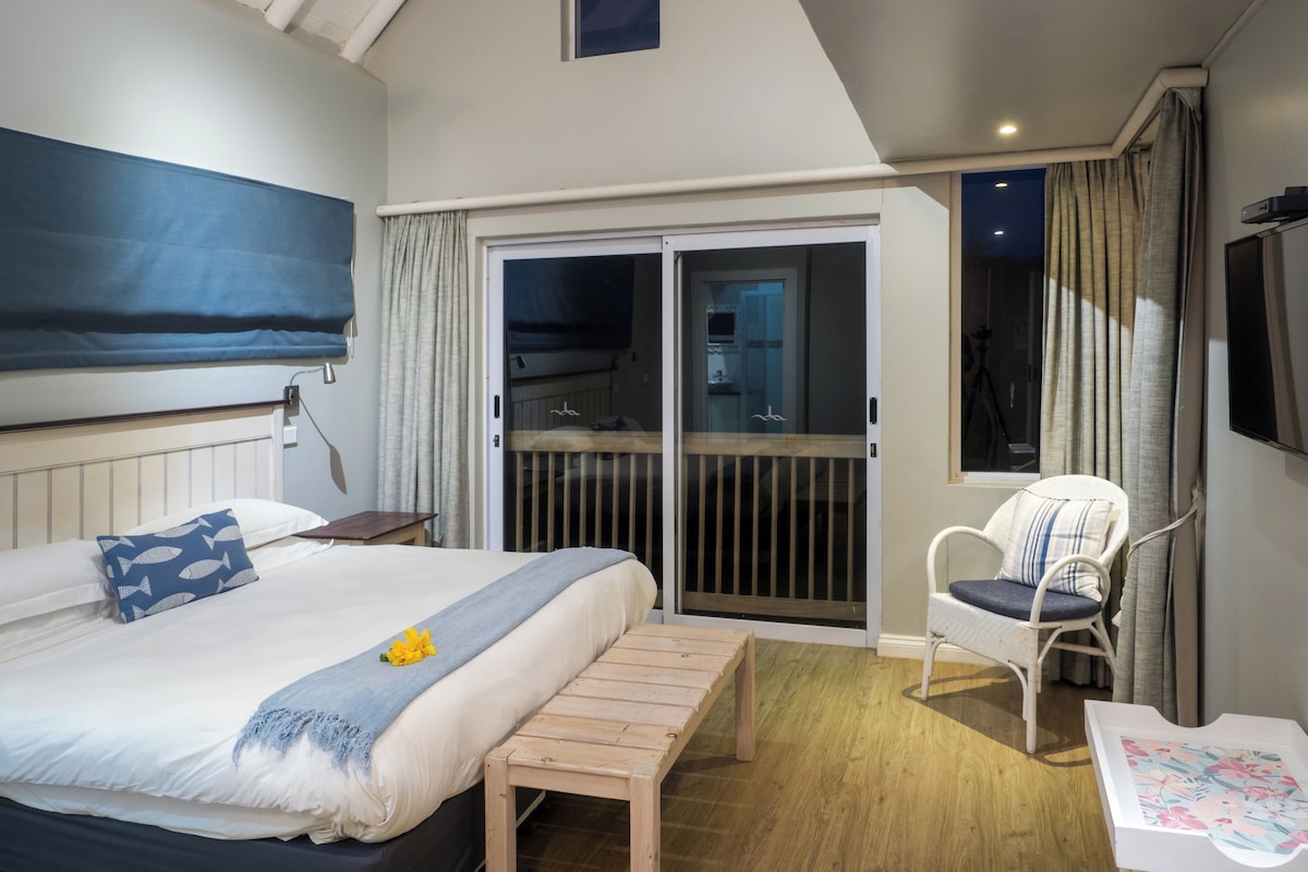 Ein Schlafzimmer mit einem Bett, einer hölzernen Bank, einem Stuhl, einem Fernseher und einer Glastür zum Balkon.