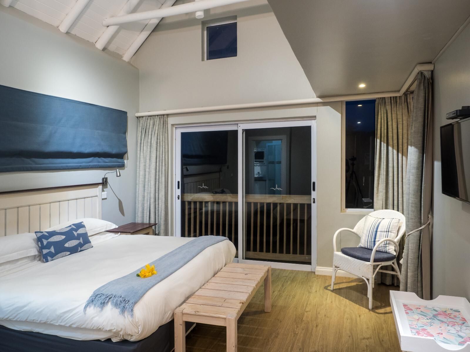 Un dormitorio con cama, un banco de madera, una silla y un ventanal corredera que da a un balcón.
