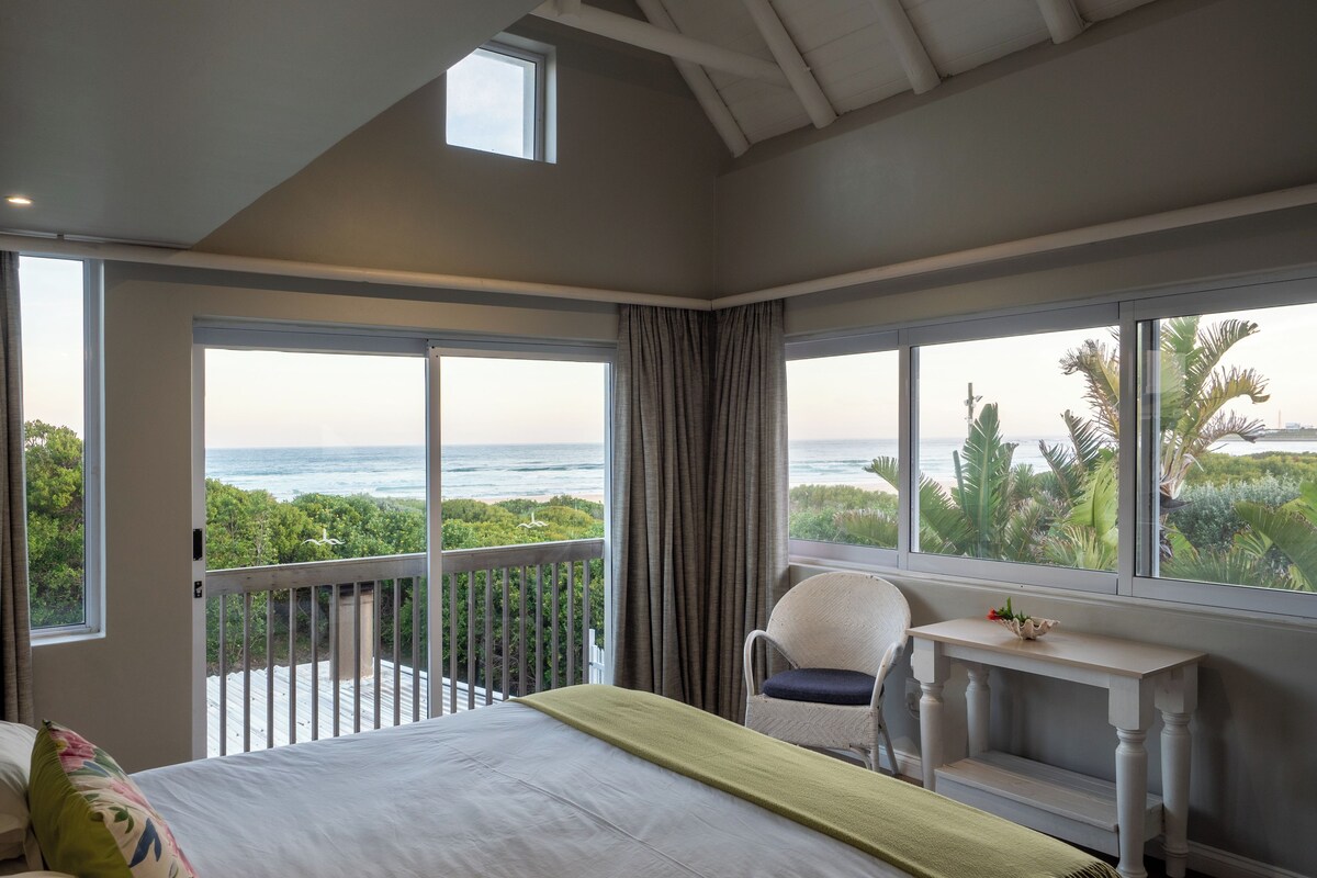 Een slaapkamer met een groot bed, een klein tafeltje, een stoel en een balkon met uitzicht op zee en groen.