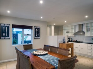 A dining area with a wooden table and wicker chairs, a kitchen with white cabinets and a stainless steel range hood, and two framed pictures on the wall. - Beach Break Guest Houses and Villas (Cape Saint Francis)