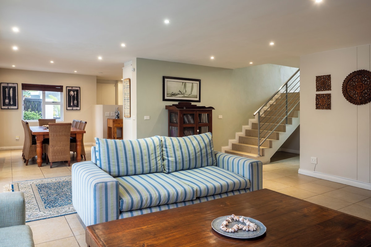 A living room with a sofa, a coffee table, a dining area with chairs, a staircase, and a bookshelf.