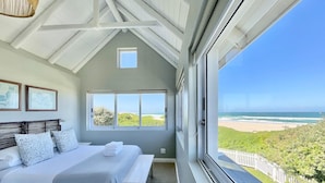 Un dormitorio con cama, un banco, un ventanal con vistas a la playa y al océano, y un techo con vigas a la vista.