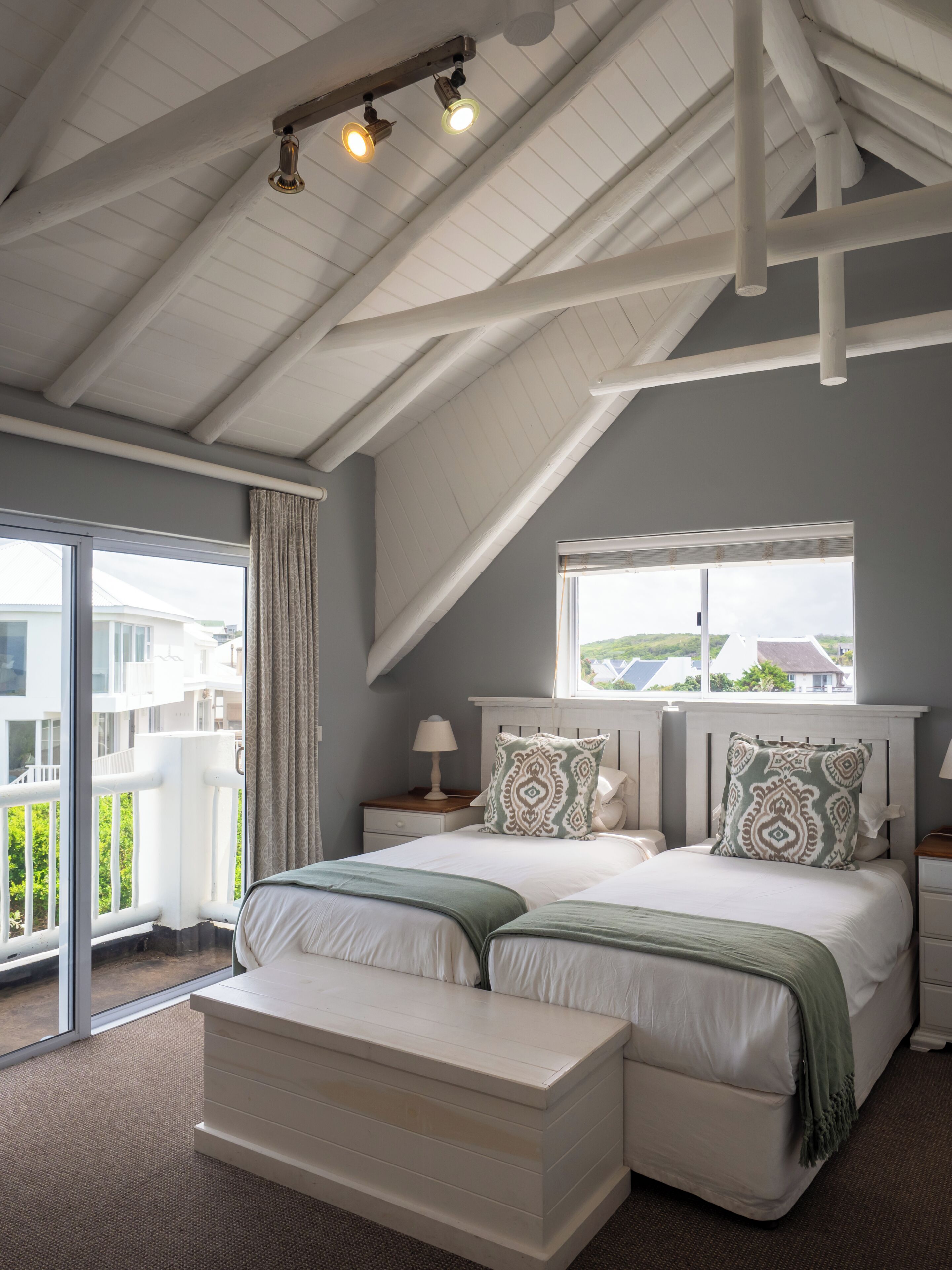 A bedroom with a sloped ceiling, a large bed with two pillows, a bedside table with a lamp, and a window with a view of buildings.