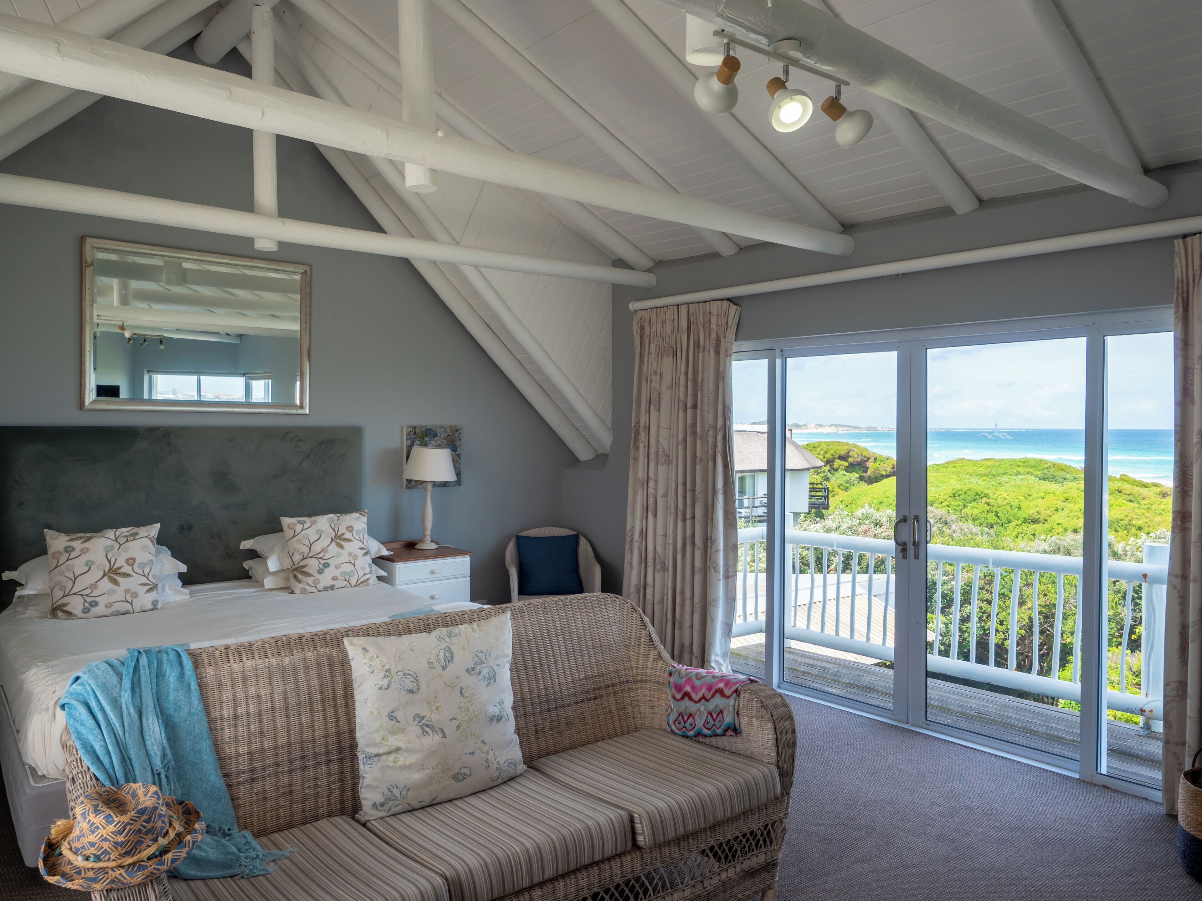 A bedroom with a bed, a sofa, a nightstand, a lamp, a mirror, and a balcony with a view of the sea.
