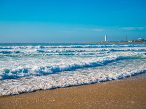 Una playa con olas, un faro y edificios a lo lejos.