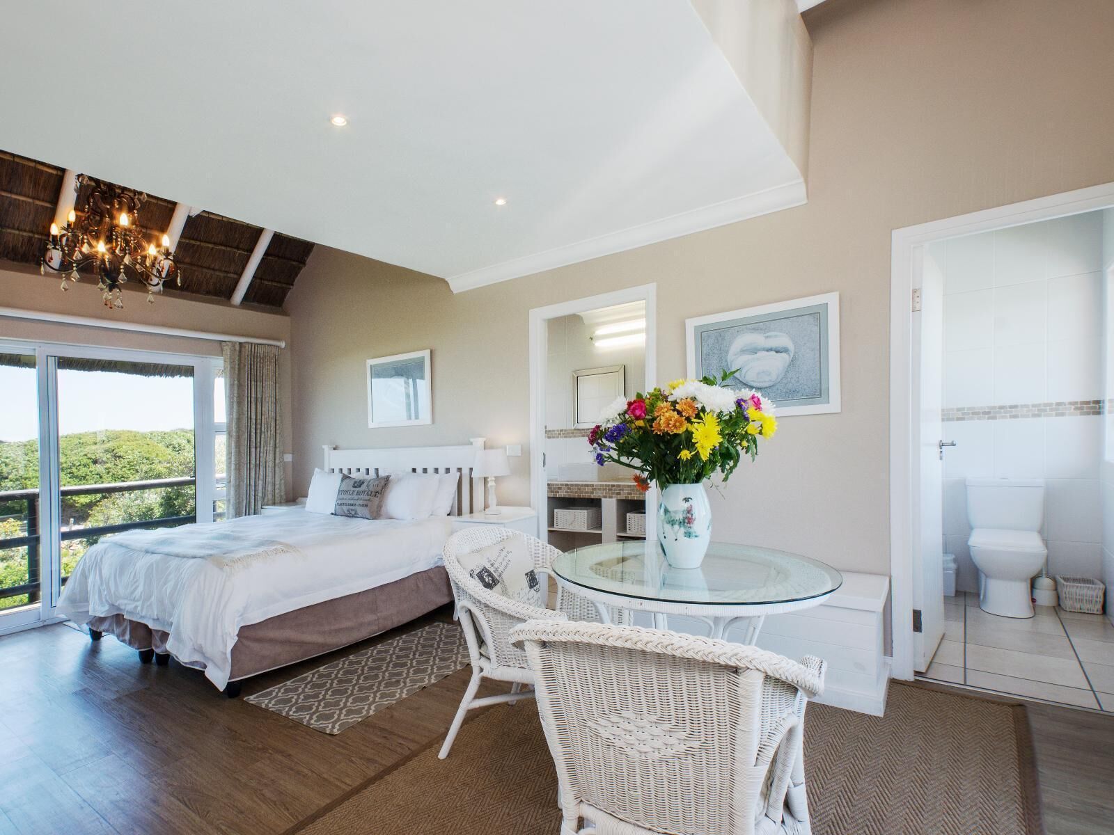 A bedroom with a large bed, a glass table, a wicker chair, and a vase of flowers.