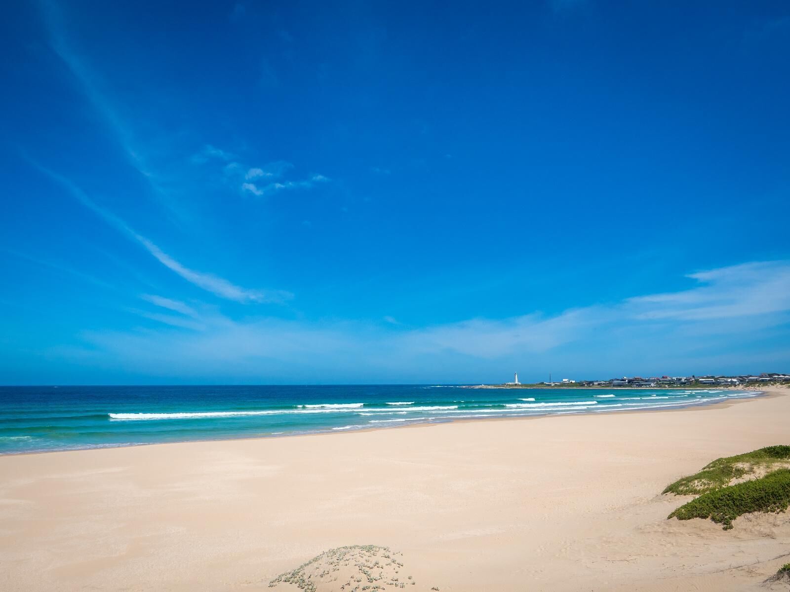 Ein Strand mit einem Leuchtturm in der Ferne, sandigem Ufer und blauem Himmel.