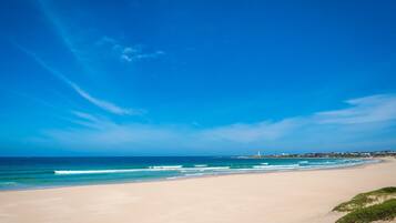 Ein Strand mit einem Leuchtturm in der Ferne, sandigem Ufer und blauem Himmel.