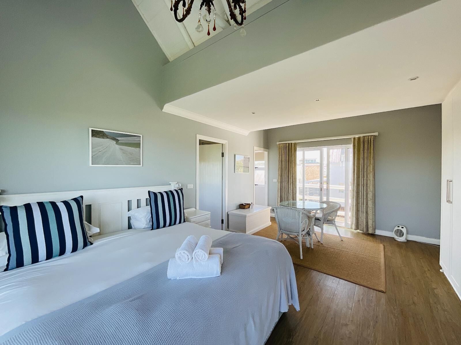 A hotel room with a large bed, a dining table with chairs, a balcony with curtains, and a ceiling fan.