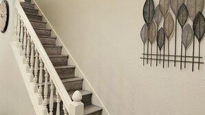 Una escalera con balaustres blancos y escalones de madera, que conduce a un rellano donde se aprecia un arreglo decorativo de hojas en la pared.