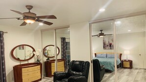 Un dormitorio con un espejo grande, un ventilador de techo, un sillón de cuero negro, un aparador de madera y una cama con colcha azul.