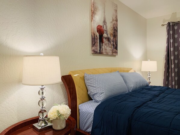A neatly arranged hotel room with a bed, two bedside lamps, a nightstand with a vase of flowers, and a painting of the Eiffel Tower.
