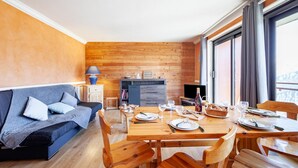 Ein gemütliches Zimmer mit Holzboden, einer Couch, einem Esstisch für vier Personen und einem Fernseher.