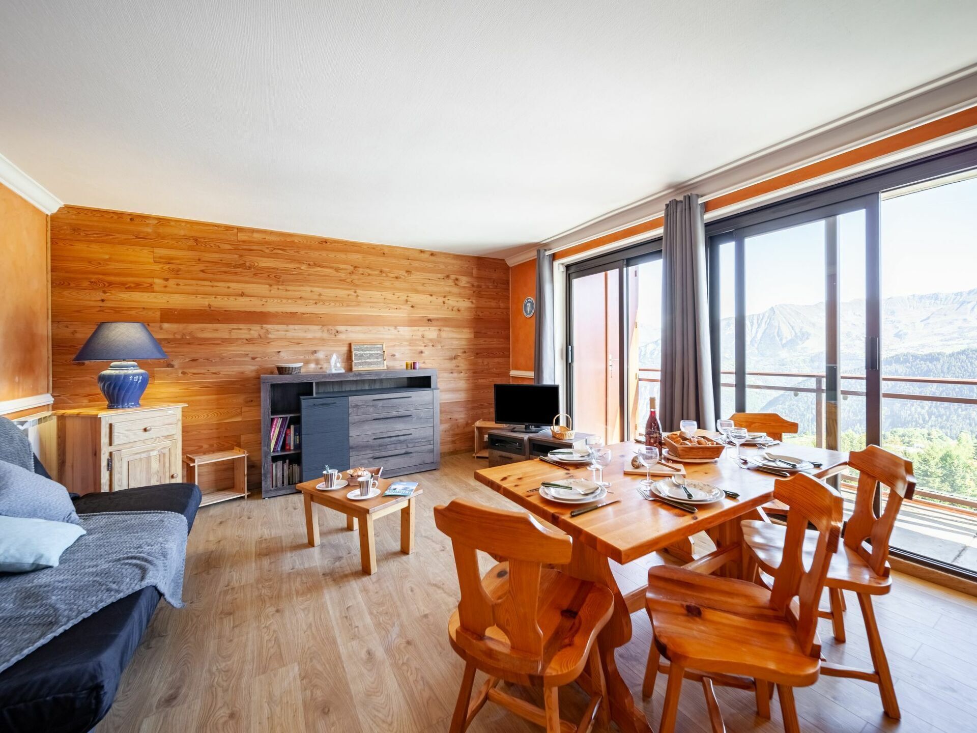 A cozy room with a wooden floor, a dining table set with plates and glasses, a sofa, a TV, and a large window offering a mountain view.