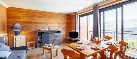A cozy room with a wooden floor, a dining table set with plates and glasses, a sofa, a TV, and a large window offering a mountain view.