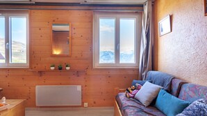 A cozy room with wooden walls, a bed with colorful bedding, a radiator, and a window with a mountain view. - Nice apartment for 4 people with WIFI, pool, TV and pets allowed (Le Corbier)