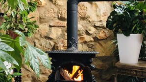 A stone fireplace with a black pipe, a fire burning inside, and a white furry rug on the floor. - House Sleeps 4 with Private Garden (Mexilhoeira Grande)