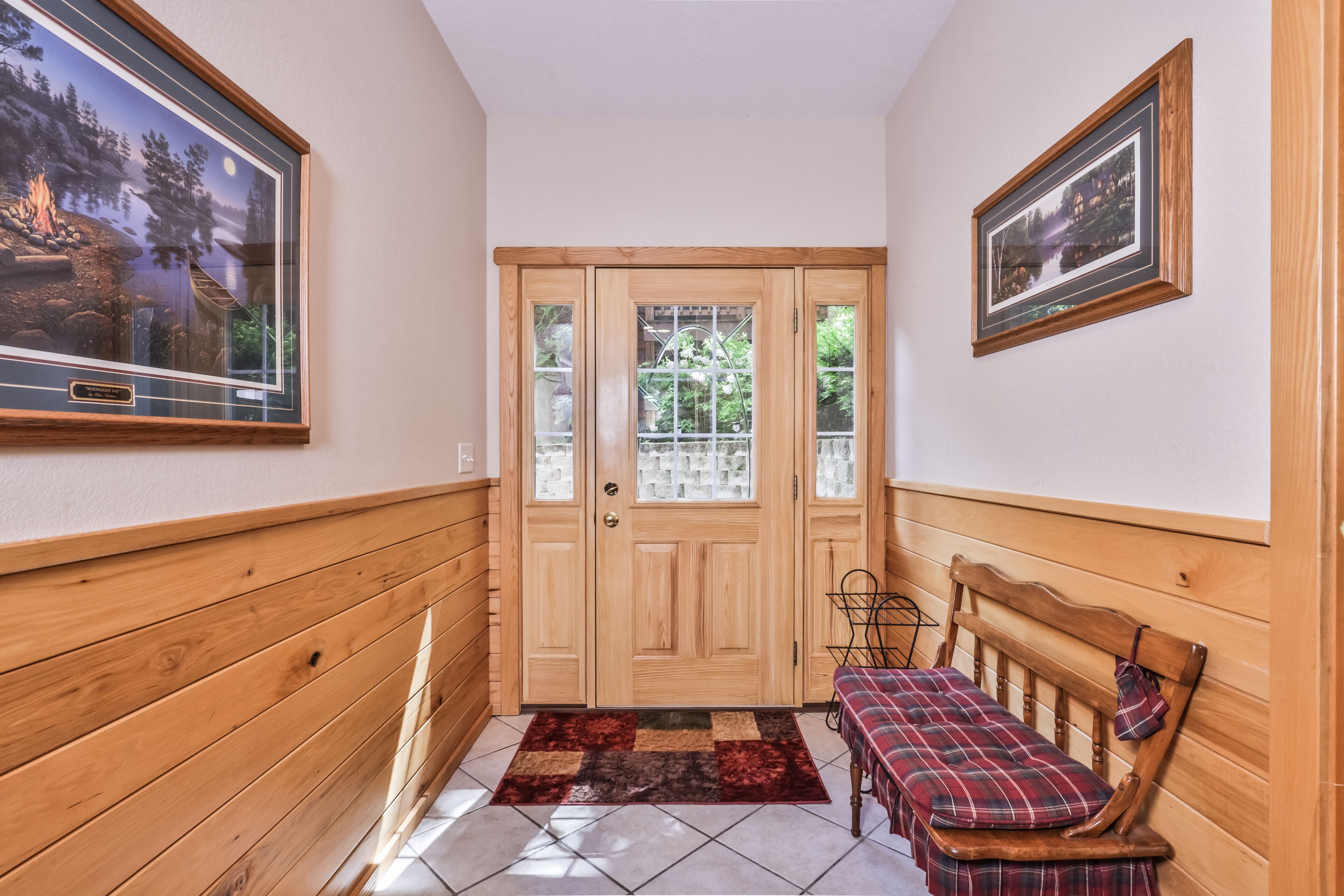 A hallway with wooden paneling, a checkered bench, a rug, and two framed pictures on the walls.