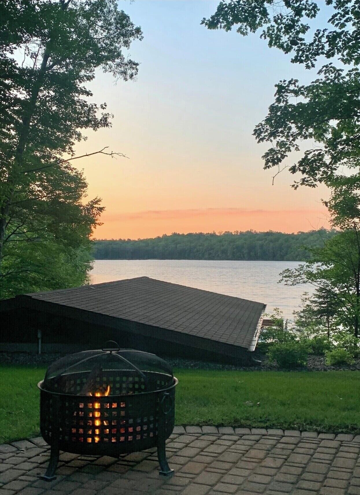 A lakeside view with a fire pit in the foreground and a building with a sloped roof in the background.