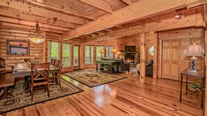 A spacious log cabin interior with a dining area, fireplace, and large windows. - Right on the river! (Sevierville)