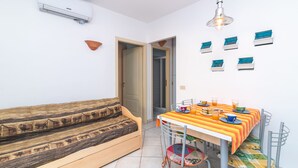 Un espacio habitable compacto con sofá, mesa de comedor y sillas. Hay un aire acondicionado montado en la pared y una lámpara colgante sobre la mesa.