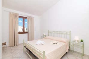 A bedroom with a bed, curtains, a window, a bedside table with a lamp, and a view of buildings outside.