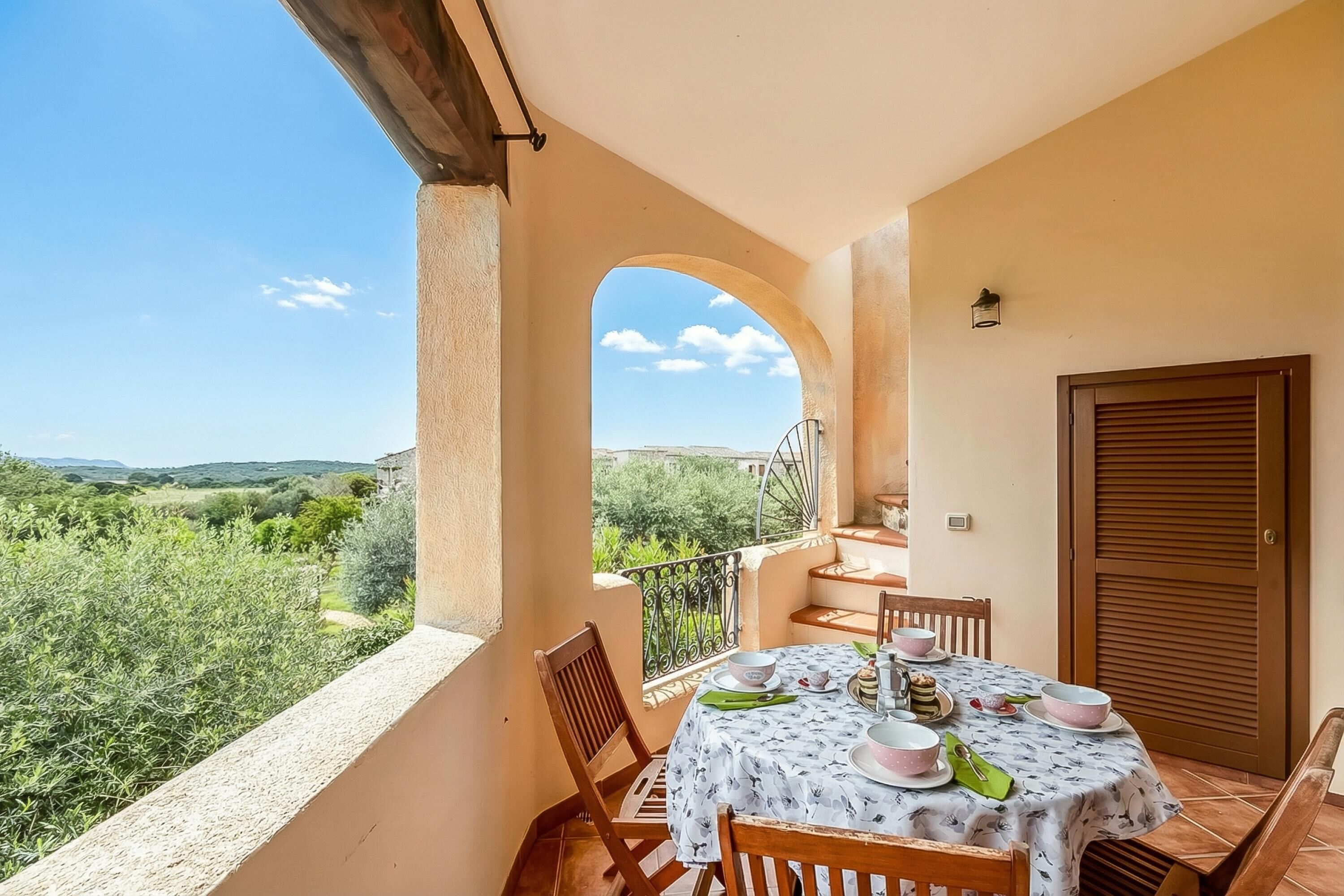 Un balcón con una mesa lista para comer, una puerta y vistas a la vegetación y colinas.