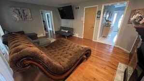A spacious living room with a large, tufted sofa, a flat-screen TV mounted on the wall, and hardwood flooring. - The Majestic Retreat Estate (West Lawn)