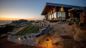 Une maison moderne dotée de grandes baies vitrées, d’une terrasse en bois et offrant une vue sur l’océan.